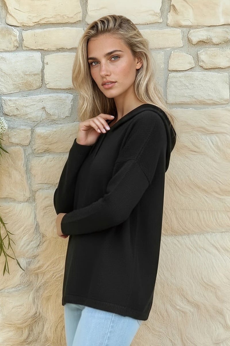 Blonde woman wearing a black hoodie, posing against a beige stone wall.