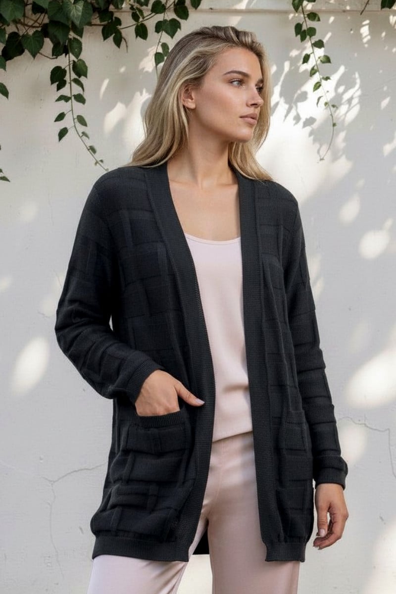 Blonde woman wearing a black cardigan with front pockets and a pale pink top, standing beside a white wall with ivy shadows.