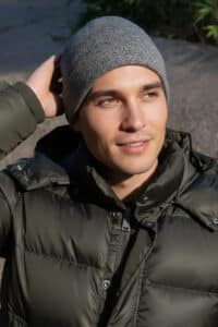 Man wearing a gray knit beanie and dark puffer jacket outdoors, looking off to the side while touching his head.